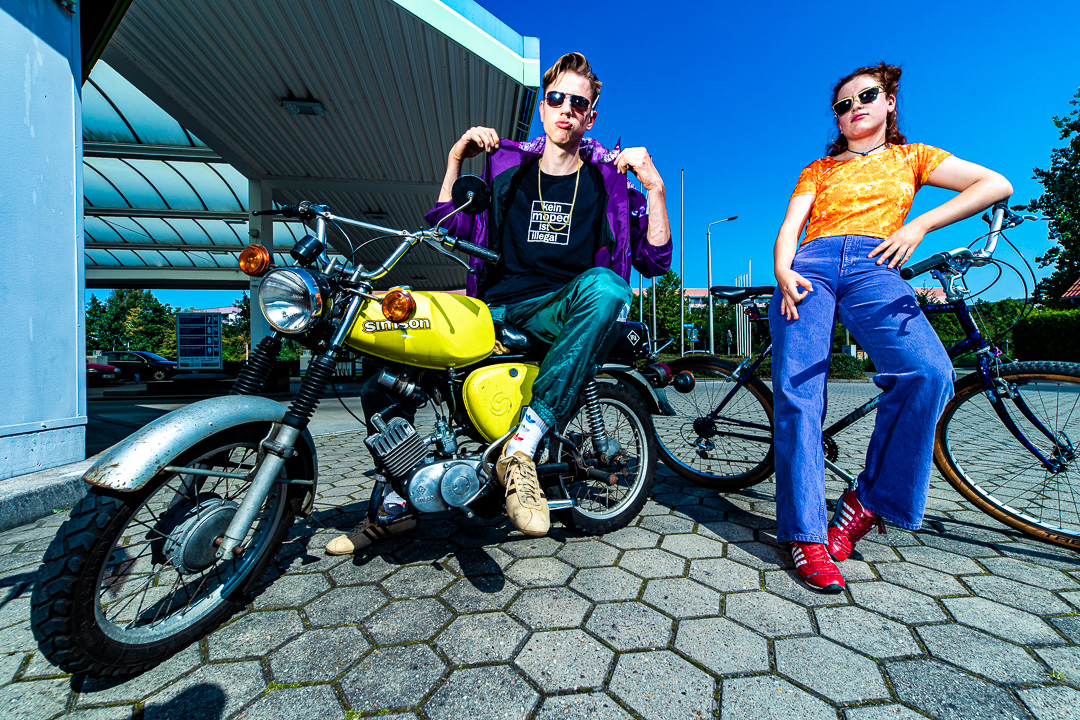 Zwei junge Erwachsene posieren selbstbewusst im Retro-Stil vor einer Tankstelle: Der Mann sitzt auf einer knallgelben Simson-Schwalbe mit der Aufschrift „Kein Moped ist illegal“, während die Frau lässig neben einem BMX-Rad steht. Beide tragen auffällige Outfits und Sonnenbrillen – urbane Szene mit Ostalgie-Flair und subkulturellem Statement