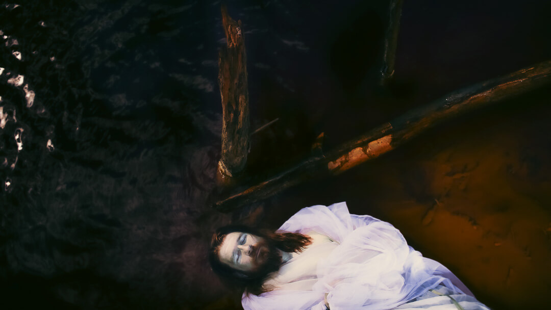 Inszeniertes Porträt einer Person mit langen Haaren in weißem Gewand, liegend im dunklen Wasser zwischen Treibholz – künstlerische Fotografie mit religiösem Motiv, aufgenommen von Fotograf Sebastian Leyser