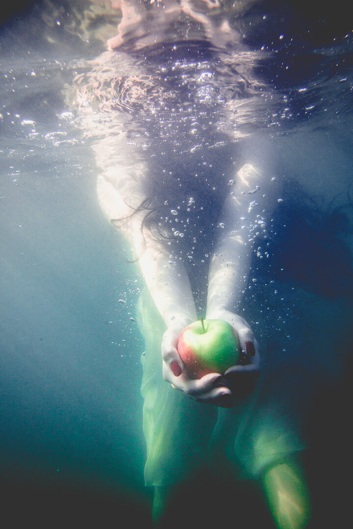 Frau unter Wasser hält einen rot-grünen Apfel mit beiden Händen in die Kamera – surrealer, symbolischer Moment in schwebender Stille.