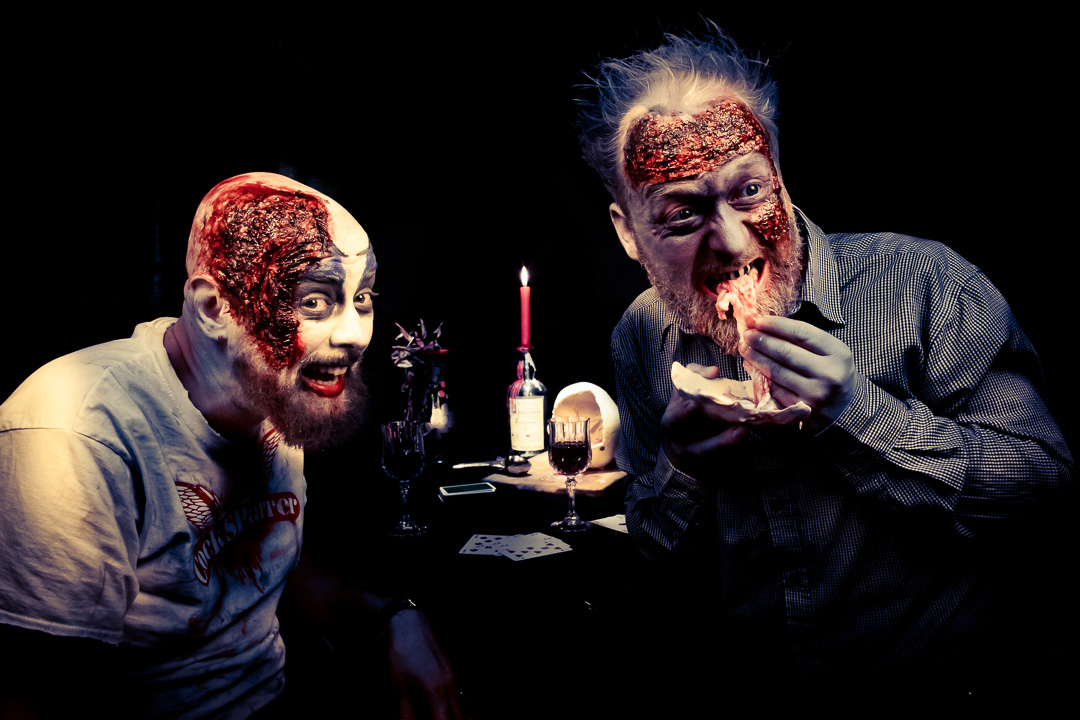 Zwei Männer mit blutigem Zombie-Make-up sitzen an einem dunklen Tisch bei Kerzenschein. Einer grinst in die Kamera, der andere beißt genüsslich in ein rohes Stück Fleisch. Horror-Szene mit makabrem Humor und starkem Kontrast – kreative Halloween-Inszenierung im Stil eines Splatter-Comedy-Shootings.