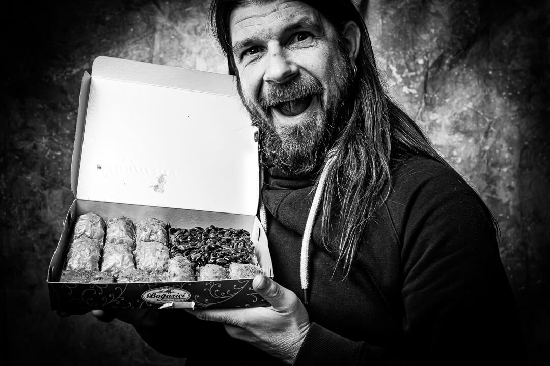 Mann mit langem Haar hält lachend eine Packung mit Baklava oder Süßgebäck in die Kamera, schwarz-weiße Studioaufnahme